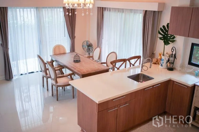 Detached House for rent in Sai Mai, Bangkok - Dining area and kitchen island in a light-filled room with sheer curtains and chandelier.