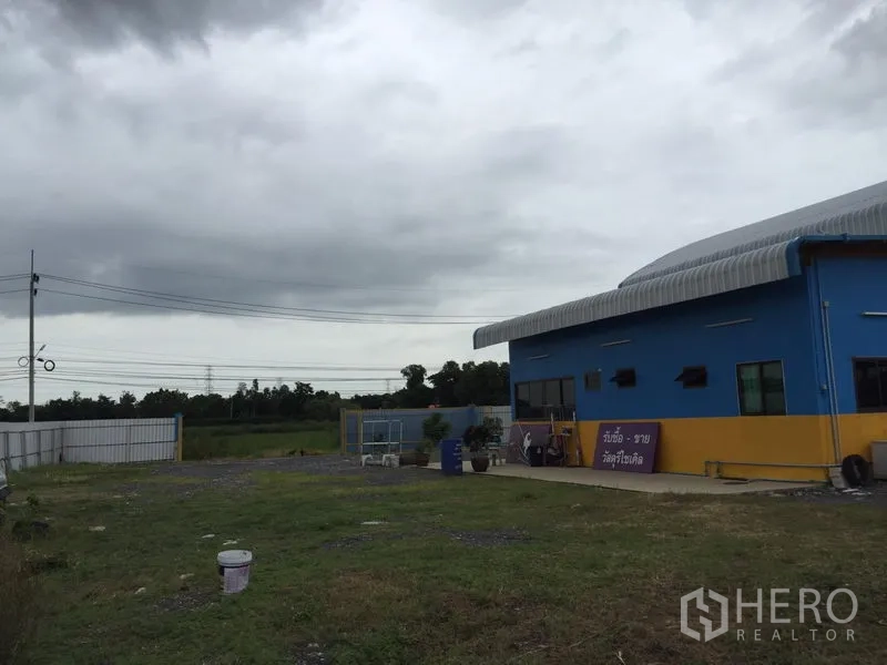Warehouse for rent in Mueang Pathum Thani, Pathum Thani - Open yard beside the warehouse with perimeter fencing and overhead utility lines.