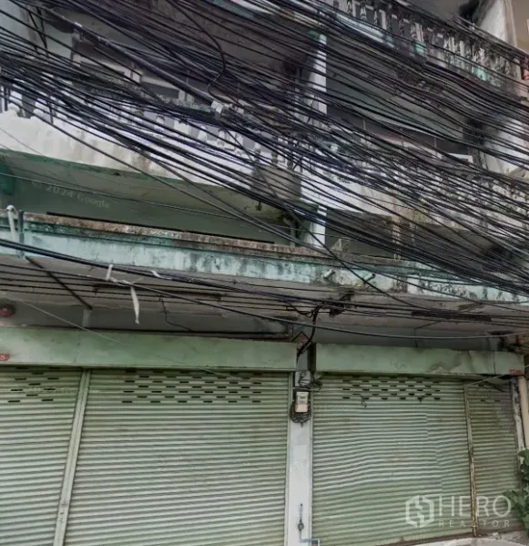 Shophouse for rent in Phra Khanong, Bangkok - Weathered multi-storey shophouse facade with closed metal roller shutters, balcony rail and dense overhead utility cables.