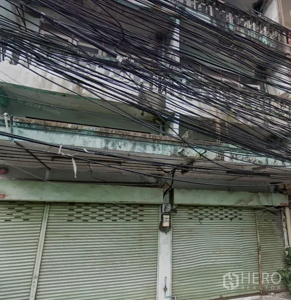 Shophouse for rent in Phra Khanong, Bangkok - Weathered multi-storey shophouse facade with closed metal roller shutters, balcony rail and dense overhead utility cables.