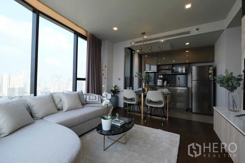 Condominium for rent in Khlong Toei, Bangkok - Bright living room with curved sofa, panoramic windows, and kitchen next to dining area.