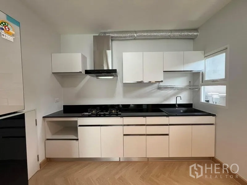 Detached House for rent in Bang Phli, Samut Prakan - White built-in kitchen with black countertops, gas hob, hood, and sink by a window.