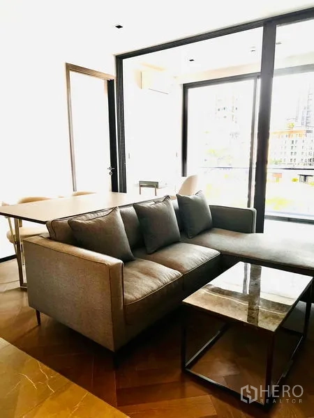 Condominium for sale in Watthana, Bangkok - Open living and dining area with gray sofa, marble‑top coffee table, dining table and full‑height windows to the balcony.