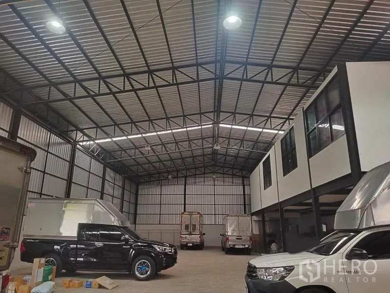 คลังสินค้า ให้เช่าในBang Khen, Bangkok - มุมมองภายในตอนค่ำ มีรถบรรทุกและออฟฟิศยกพื้นใต้โครงหลังคาเหล็ก.