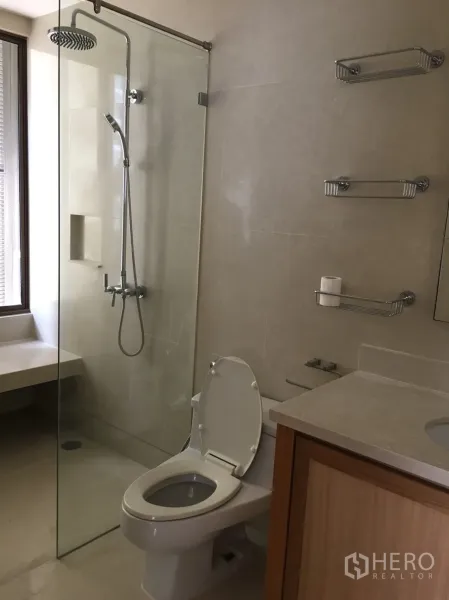 公寓 出租在Khlong Toei, Bangkok - 现代浴室，玻璃淋浴间与顶喷花洒，墙面置物架和浅色木质洗手台。