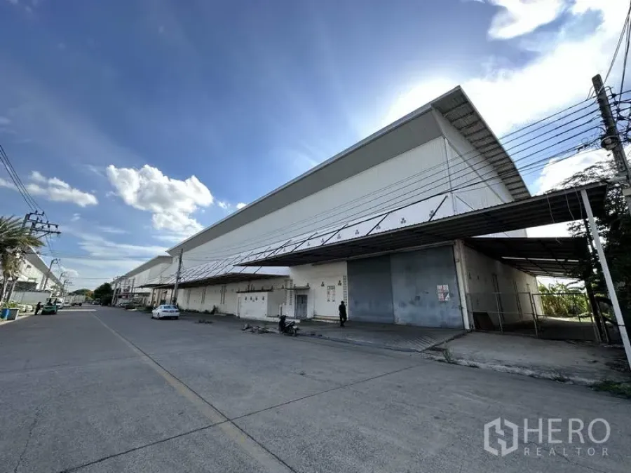 Factory for rent in Bang Bua Thong, Nonthaburi - Street view of the long factory facade with metal doors and awnings under a blue sky.