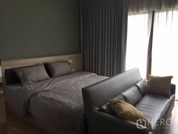 Condominium for rent or sale in Bangkok - Cozy bedroom with queen bed, grey curtains, balcony sliding doors and a leather sofa at the foot of the bed.
