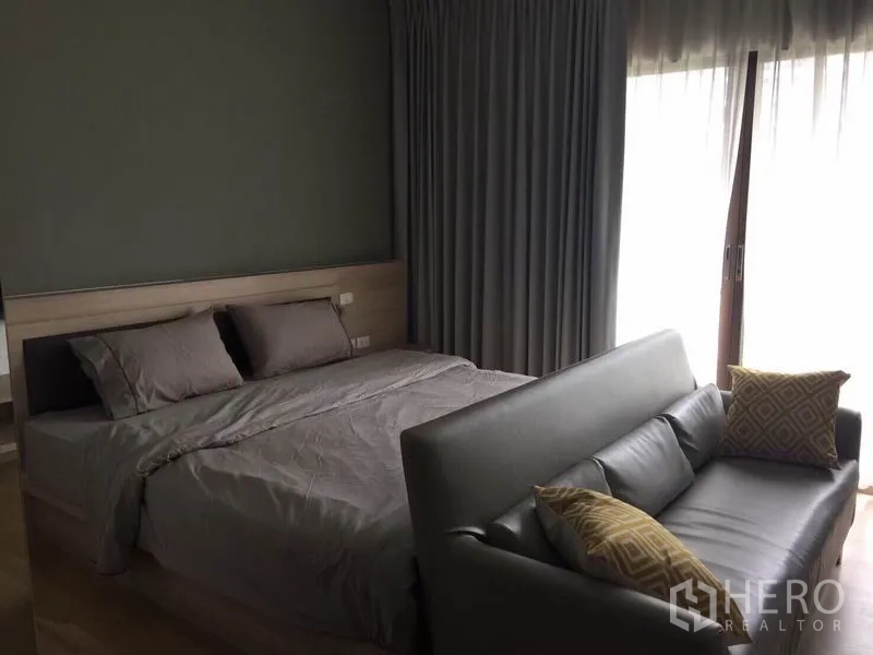 Condominium for rent or sale in Bangkok - Cozy bedroom with queen bed, grey curtains, balcony sliding doors and a leather sofa at the foot of the bed.