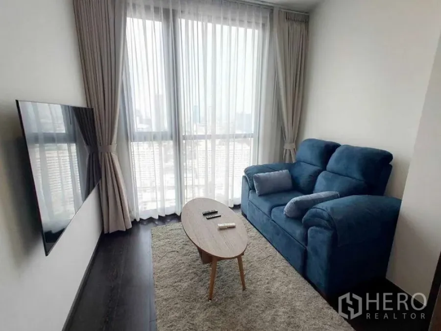 Condominium for rent in Watthana, Bangkok - Living room with a blue sofa, wall-mounted TV, and floor-to-ceiling windows.