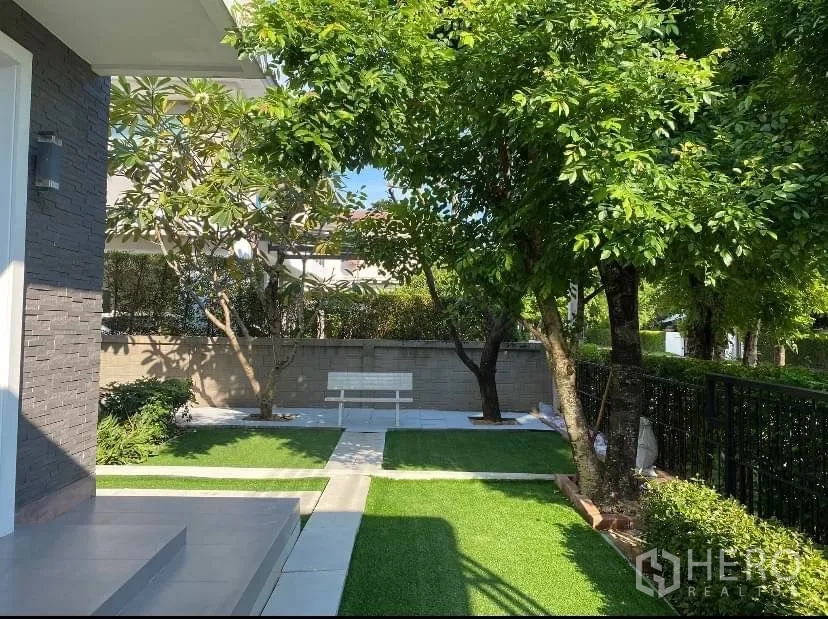 Detached House for rent or sale in Prawet, Bangkok - Landscaped front garden featuring mature shade trees, a bench and paved walkways.