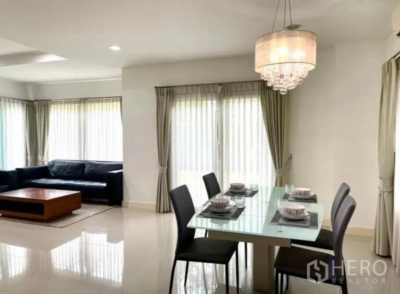 Detached House for rent or sale in Lat Krabang, Bangkok - Dining area with glass table, four chairs and a chandelier next to full-height windows.