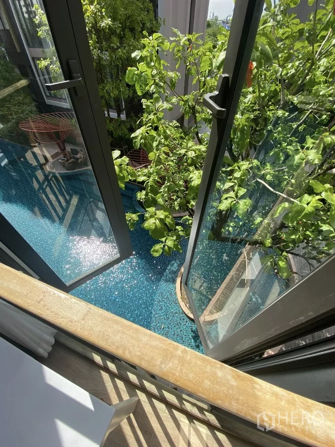 Condominium for rent or sale in Watthana, Bangkok - Open balcony doors overlooking a turquoise mosaic swimming pool framed by lush garden trees.