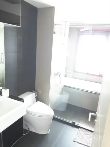 Condominium for rent in Khlong Toei, Bangkok - Modern bathroom with glass-enclosed shower, toilet, and vanity beside a window.