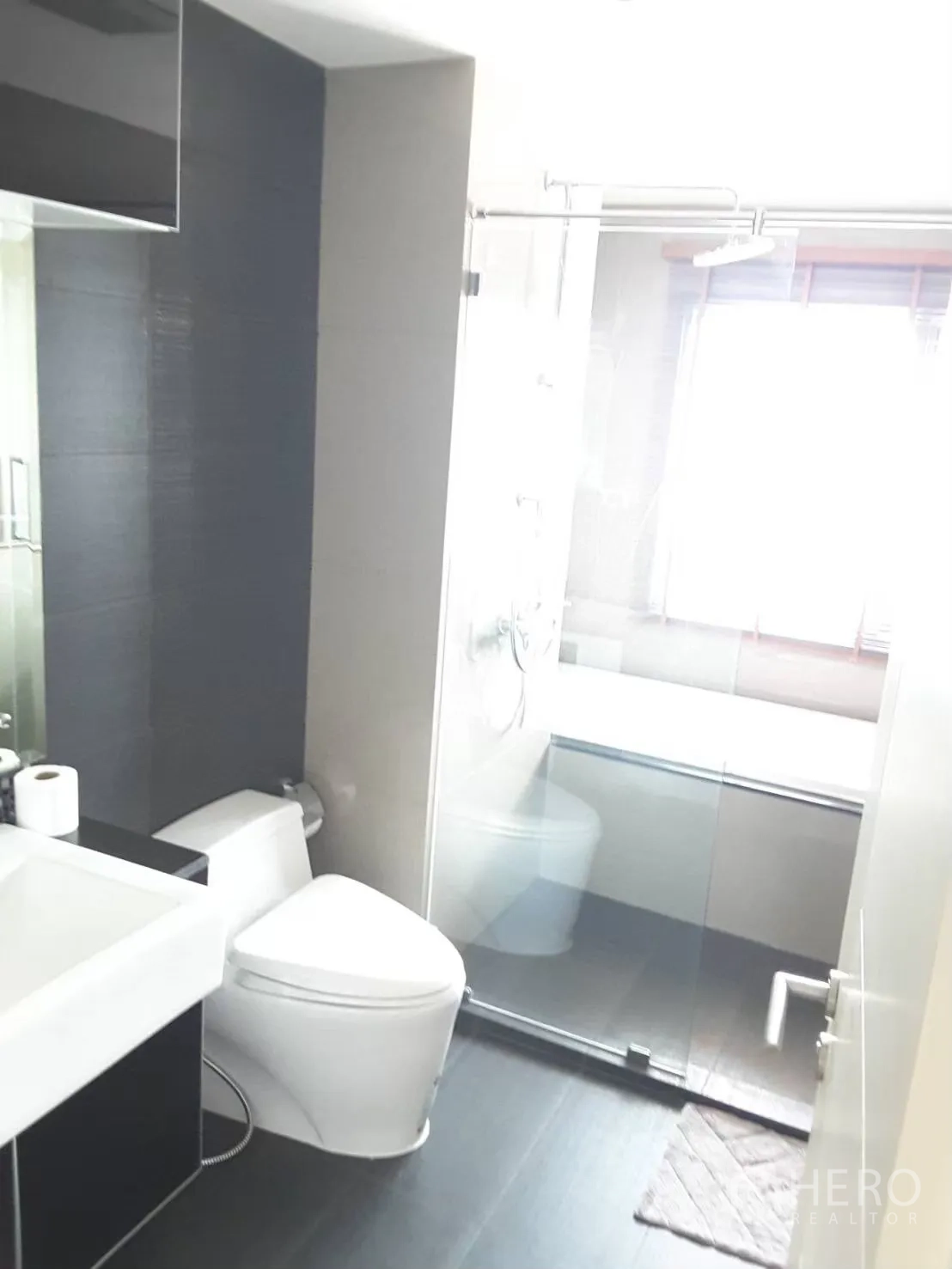 Condominium for rent in Khlong Toei, Bangkok - Modern bathroom with glass-enclosed shower, toilet, and vanity beside a window.