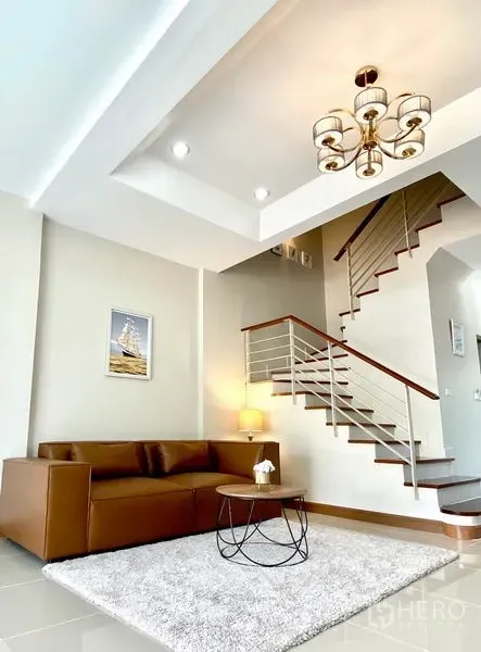 Townhouse for rent in Prawet, Bangkok - Cozy lounge corner with brown leather sofa, rug and sleek open staircase under tray ceiling.