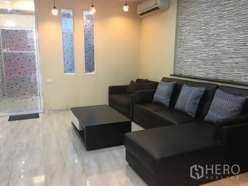 联排别墅 出租在Bang Rak, Bangkok - 温馨客厅，深色皮沙发、茶几、吊灯、空调与磨砂玻璃门，瓷砖地面。