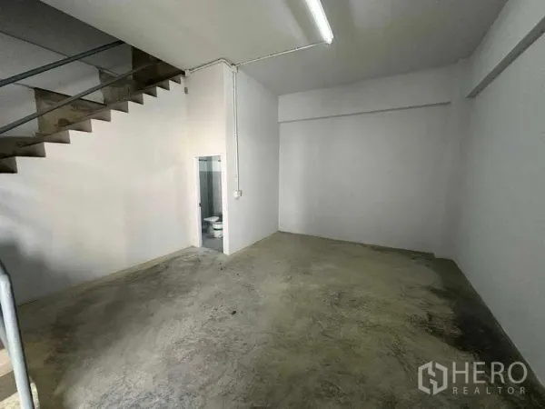 货栈 出租在Samut Sakhon - 一层工作区，水泥地面，楼梯通往上层，旁有卫生间门。