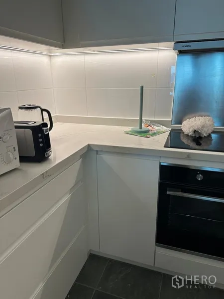 Condominium for rent in Khlong Toei, Bangkok - Compact L-shaped kitchen with quartz counters, built-in oven, and small appliances.