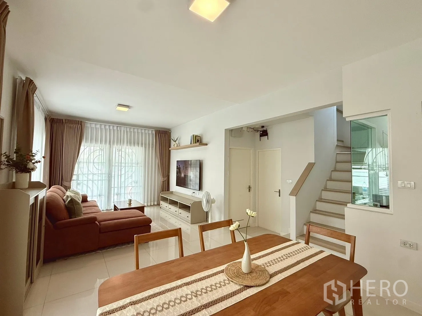 Detached House for rent in Bang Phli, Samut Prakan - Dining table facing a cozy lounge and staircase with white tile floors and curtains.