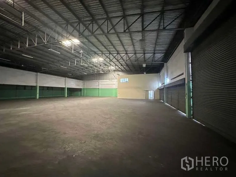Factory for rent in Nonthaburi - Large warehouse hall with high steel-truss roof, concrete floor and multiple roller shutter doors.