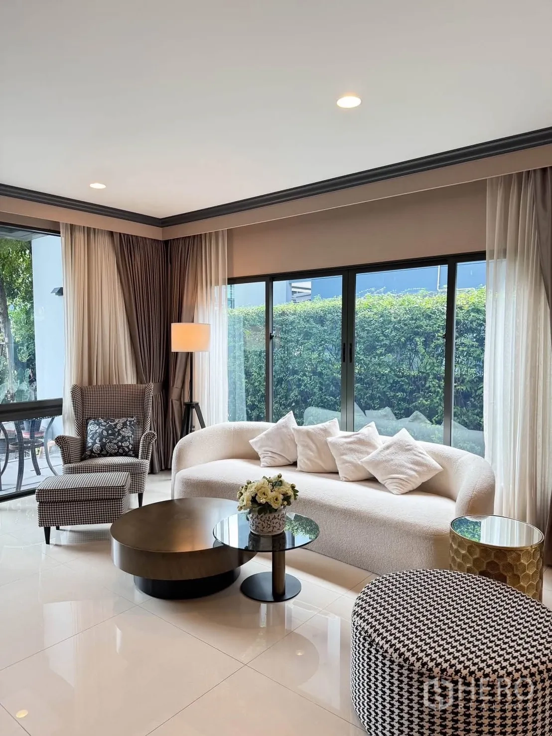 Detached House for rent in Bang Kapi, Bangkok - Bright lounge with curved white sofa, houndstooth ottoman, and garden view through large windows.