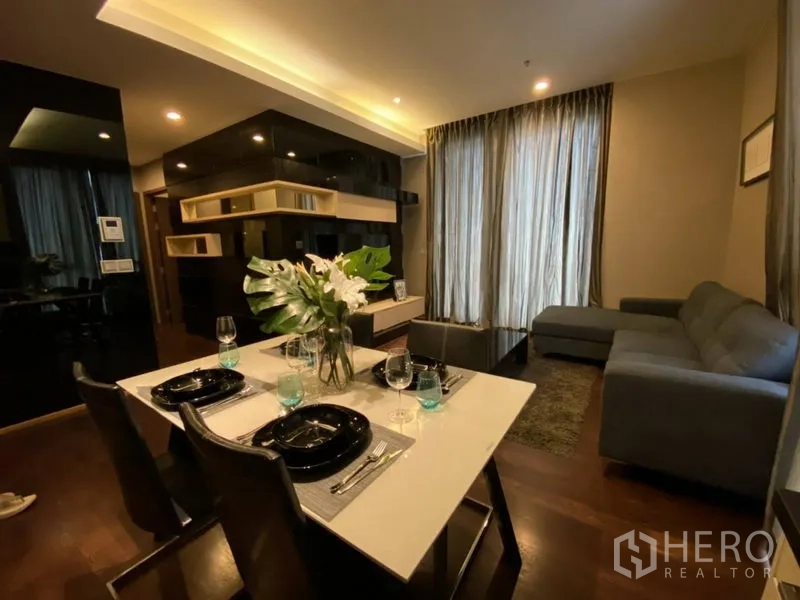 Condominium for rent in Watthana, Bangkok - Open-plan living and dining room with L-shaped sofa, built-in shelving, wooden floors and full-height curtains in a Thonglor condo.