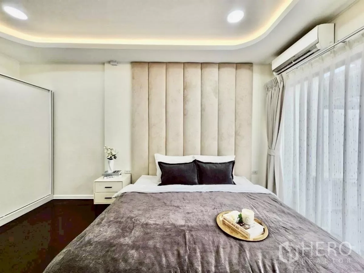 Townhouse for rent in Huai Khwang, Bangkok - Primary bedroom with padded headboard, soft recessed lighting and large curtained window.