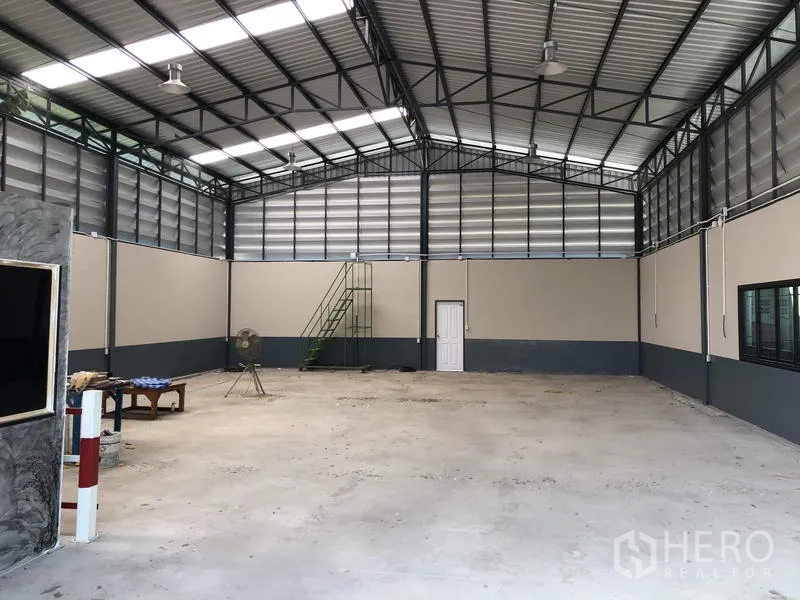 货栈 出售在Pathum Thani - 库内广角视图，通风板、吊灯、窗户及一侧绿色金属楼梯。