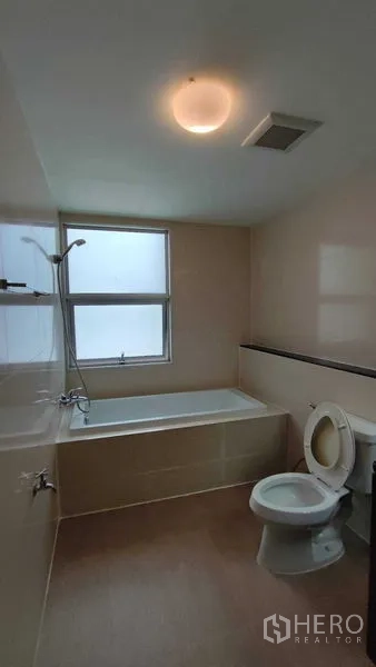 Condominium for rent in Khlong Toei, Bangkok - Bathroom with bathtub beneath a frosted window, overhead shower and toilet.