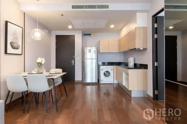 Condominium for rent in Pathum Wan, Bangkok - Kitchenette with light wood cabinets, black countertops, fridge, washing machine, and a four-seat dining set.