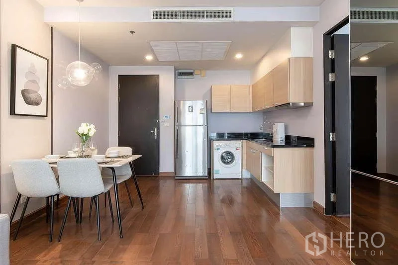 Condominium for rent in Pathum Wan, Bangkok - Kitchenette with light wood cabinets, black countertops, fridge, washing machine, and a four-seat dining set.