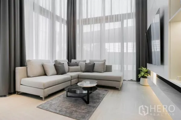 Townhouse for sale in Chatuchak, Bangkok - Double-volume living room with L-shaped sofa, sheer curtains and wall-mounted TV.