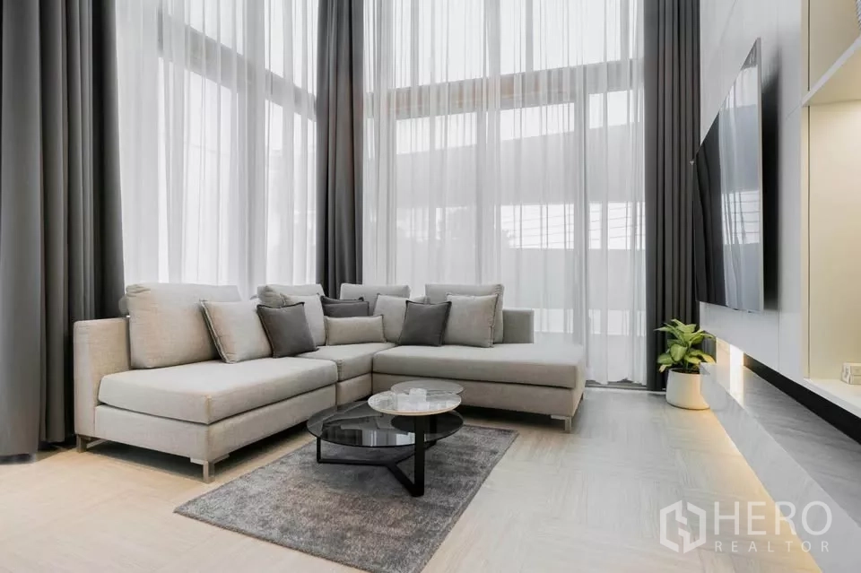 Townhouse for sale in Chatuchak, Bangkok - Double-volume living room with L-shaped sofa, sheer curtains and wall-mounted TV.