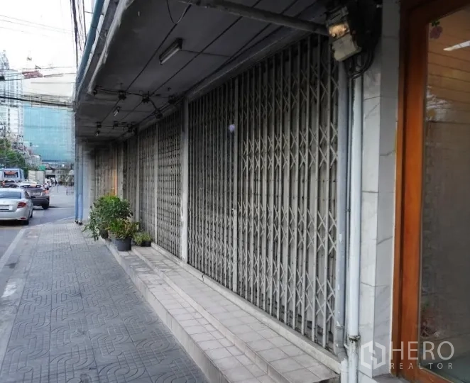 Shophouse for rent in Khlong Toei, Bangkok - Covered sidewalk with closed accordion metal shopfront gates and street traffic in front of a row of shophouses.