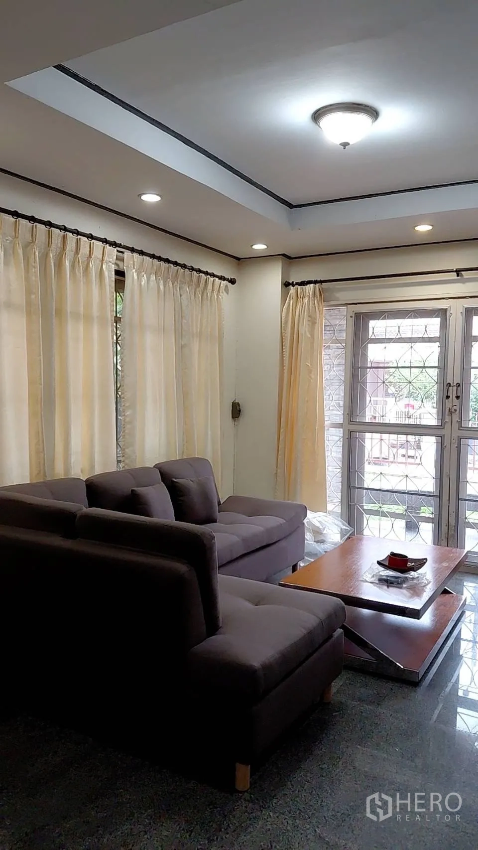 独立式住宅 出租在Min Buri, Bangkok - 起居区转角沙发、浅色窗帘与通向前庭的玻璃门。