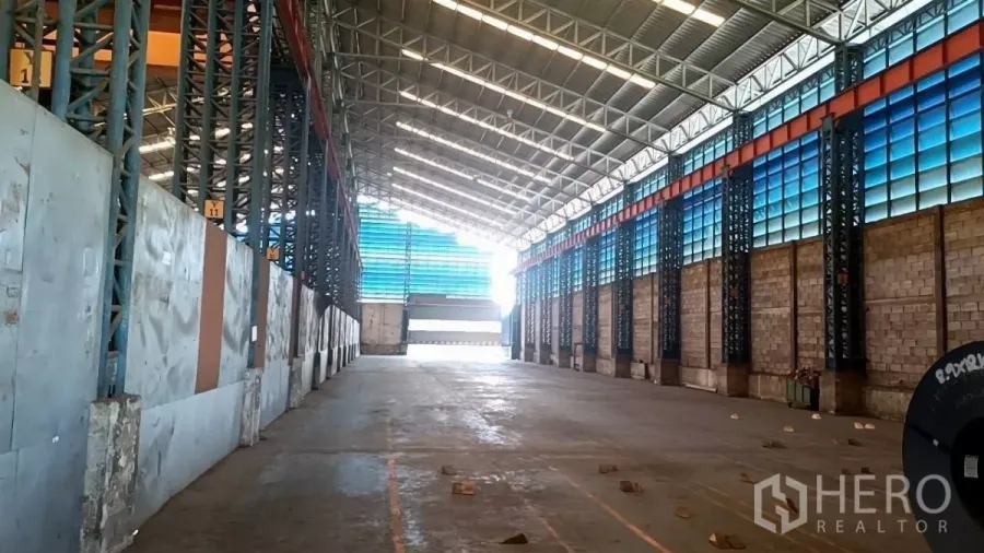 Warehouse for rent in Mueang Samut Sakhon, Samut Sakhon - Spacious warehouse bay with high steel truss roof, blue translucent panels, and a wide loading door on reinforced concrete.