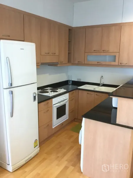 Condominium for rent in Khlong Toei, Bangkok - L-shaped kitchen with wood cabinetry, black stone countertop, white oven, cooktop and refrigerator.