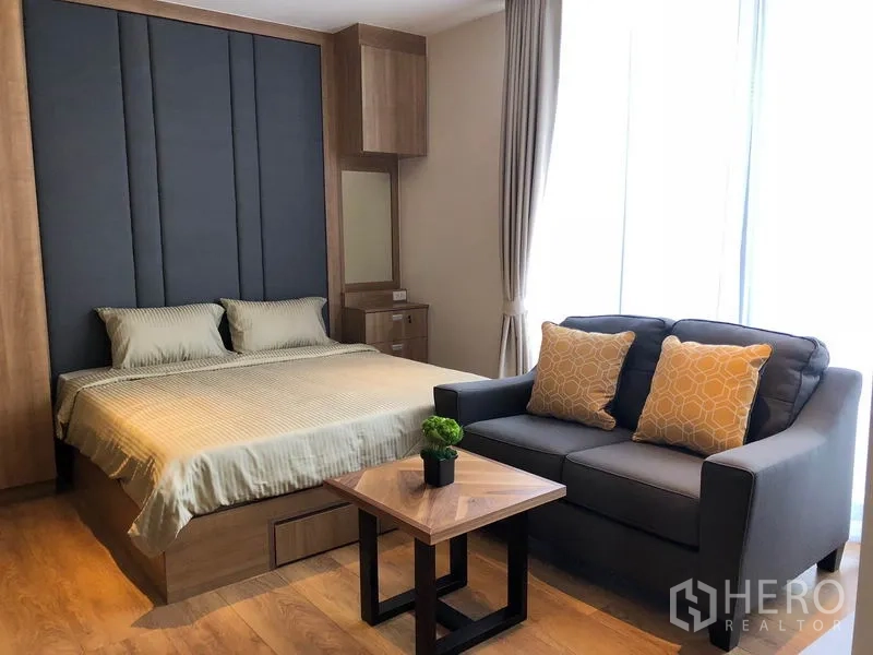 Condominium for rent in Khlong Toei, Bangkok - Cozy sleeping area with tall upholstered headboard, gray sofa with yellow cushions and coffee table.