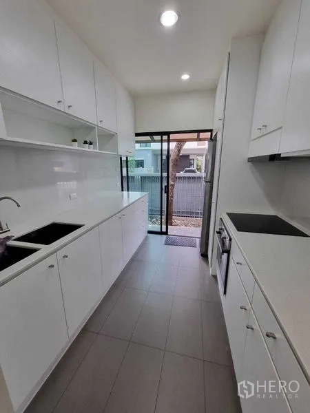 Detached House for rent or sale in Prawet, Bangkok - Long white galley kitchen with cabinets, sink, cooktop and door to the yard.