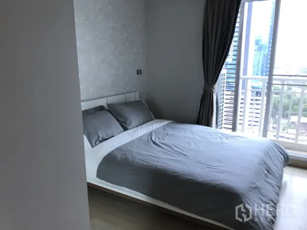 公寓 出租在Watthana, Bangkok - 次卧铺灰色床品，落地窗与窗帘，小阳台可见城市天际线。