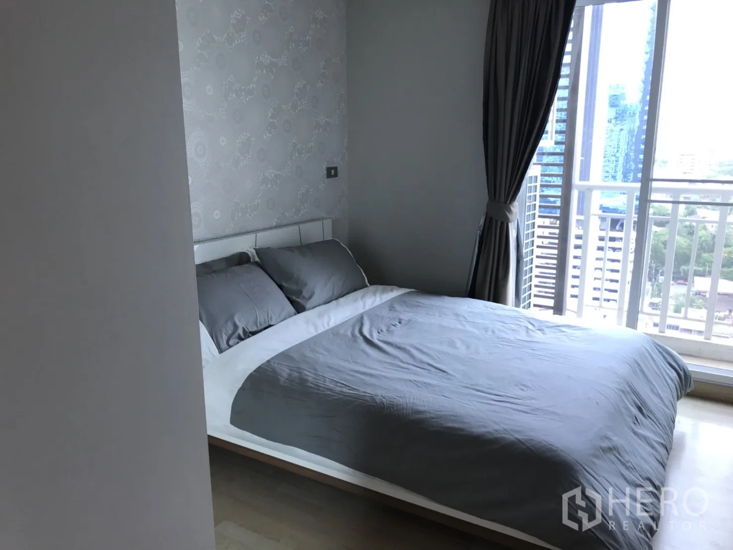 公寓 出租在Watthana, Bangkok - 次卧铺灰色床品，落地窗与窗帘，小阳台可见城市天际线。