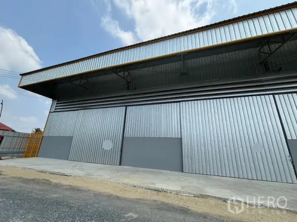 Warehouse for rent in Mueang Pathum Thani, Pathum Thani - Front exterior of a metal-clad warehouse with wide sliding doors, deep canopy and loading apron under a blue sky.