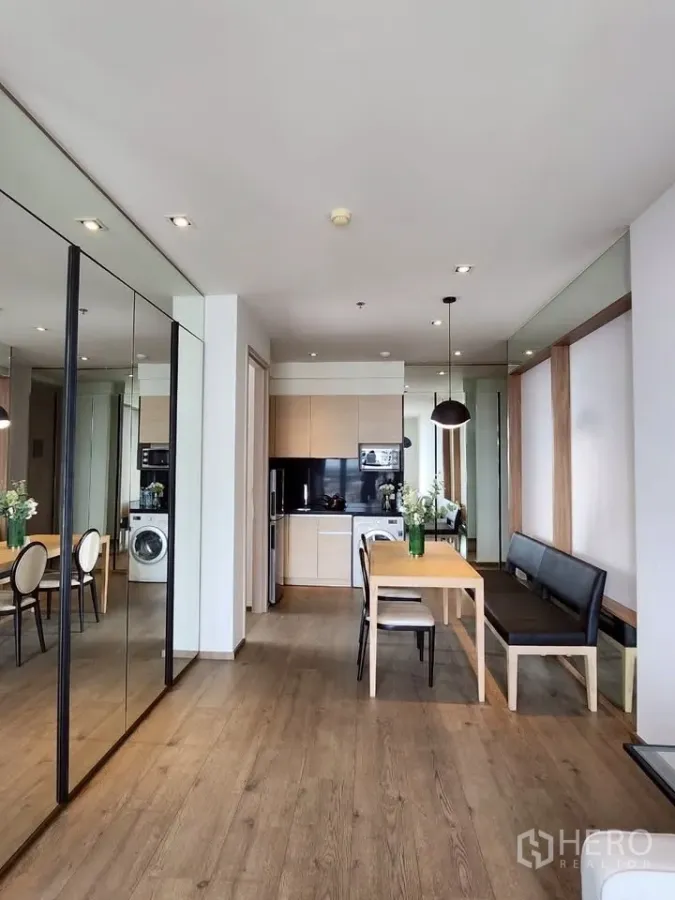 Condominium for rent in Khlong Toei, Bangkok - Spacious dining area with mirrored wall, warm wood flooring and a compact kitchen with in‑unit washing machine.