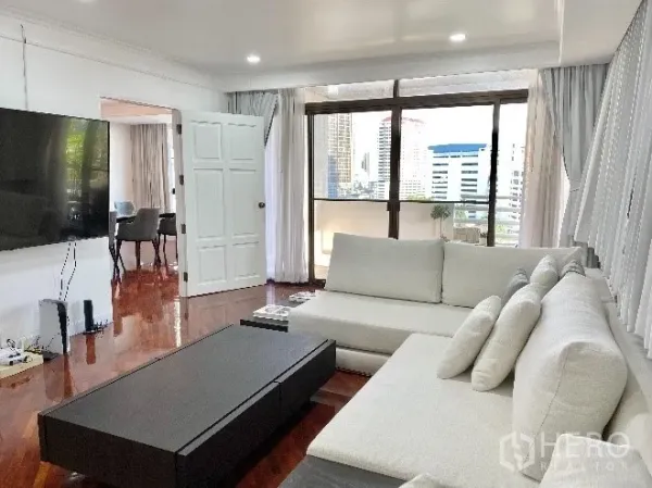 Condominium for rent in Khlong Toei, Bangkok - Lounge with large sectional sofa, wall-mounted TV and floor-to-ceiling windows leading to a balcony with city views.