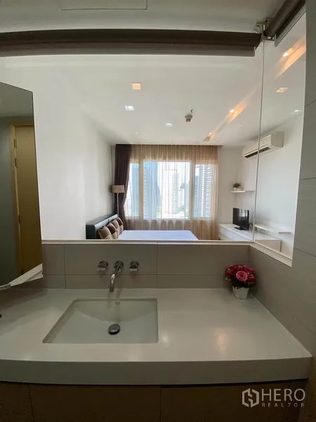 Condominium for rent in Khlong Toei, Bangkok - Contemporary bathroom sink area overlooking a bright bedroom through a glass wall.