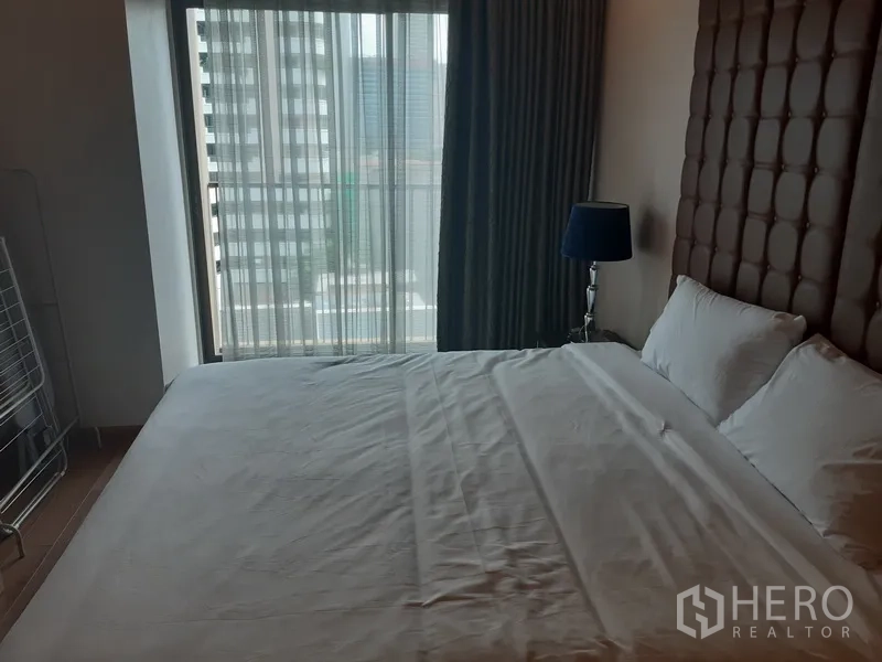 公寓 出租在Bangkok - 卧室配特大床、软包床头板，落地窗与窗帘可观城市景色。