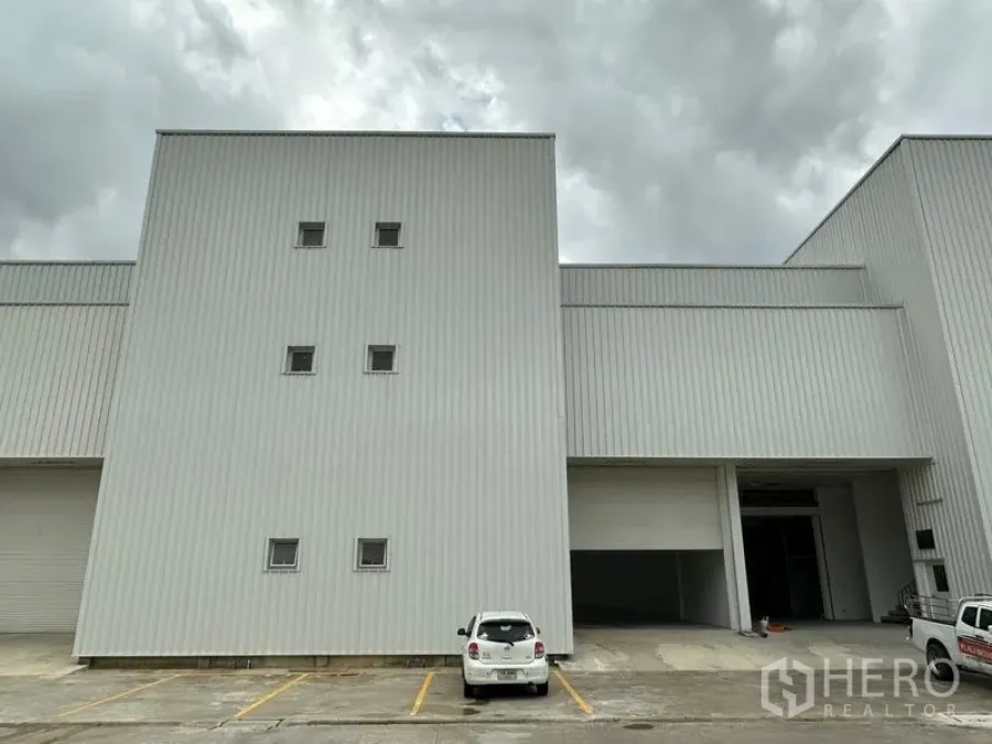 Factory for rent in Khlong Luang, Pathum Thani - Front elevation of a modern white factory with high roller doors and marked parking bays under a cloudy sky