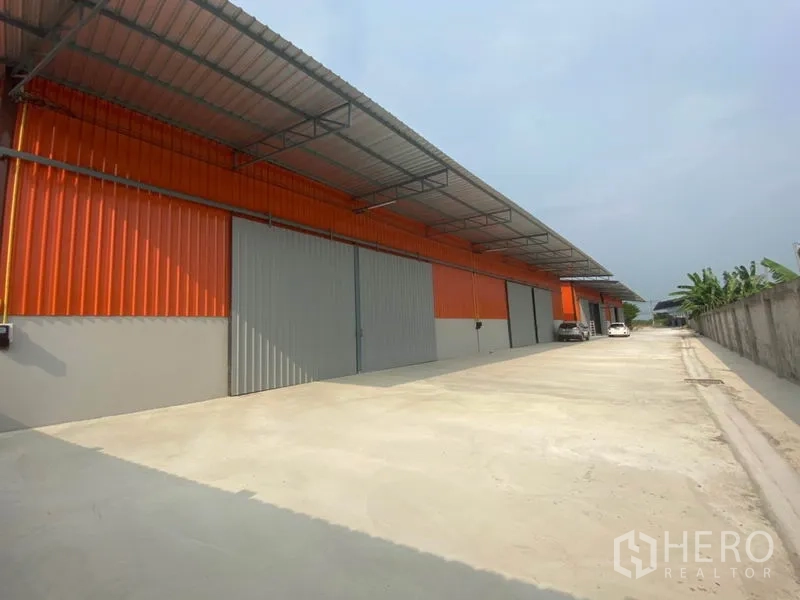 Warehouse for rent in Bang Yai, Nonthaburi - Orange metal warehouse facade with large grey sliding doors and a spacious truck-friendly driveway.