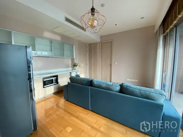Condominium for rent or sale in Yan Nawa, Bangkok - Living area facing kitchenette and hallway with a pendant light and a large blue sectional sofa on polished wood floors.