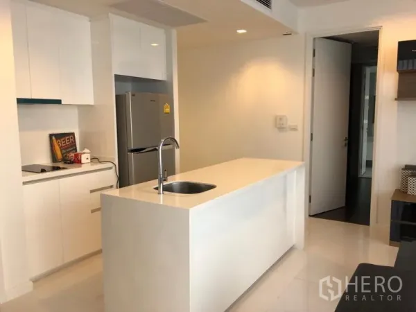 Condominium for rent in Sathon, Bangkok - Modern white kitchen with a long island and sink, tall refrigerator, built‑in cooktop and sleek cabinetry.